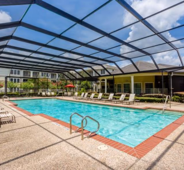 Covered outdoor swimming pool with lounge chairs and surrounding apartment buildings under a large screened enclosure.