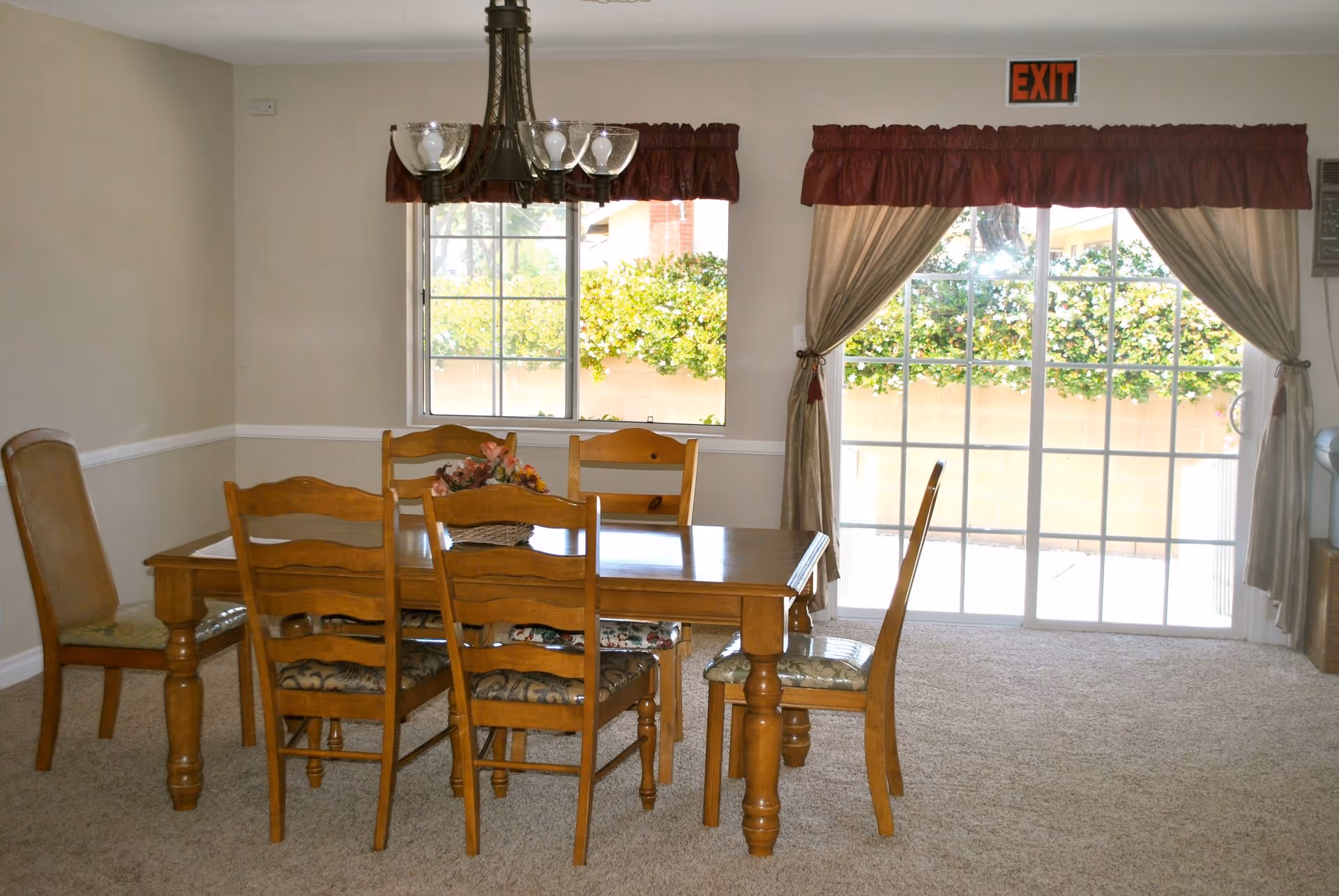 A dining room with a wooden dining table and six chairs, a floral centerpiece on the table, beige carpeted floor, two windows with curtains, and an exit sign above a sliding glass door leading outside.