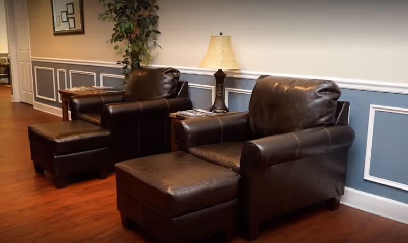 Two dark brown leather armchairs with matching ottomans are placed on a wooden floor against a wall with blue and white wainscoting. A small wooden side table with a decorative lamp and some magazines is positioned between the chairs. A tall green plant is visible in the background near a doorway.