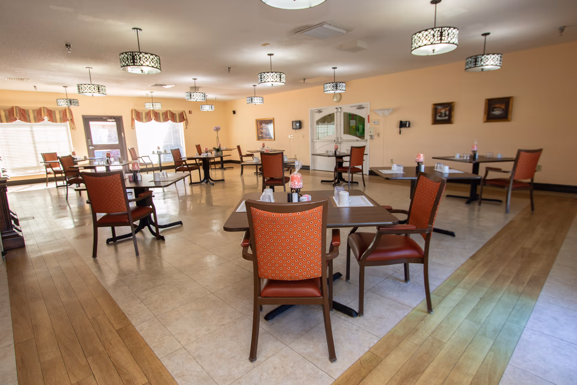 Spacious dining room with multiple tables and red-upholstered chairs, pendant lights, and windows in a senior living facility.