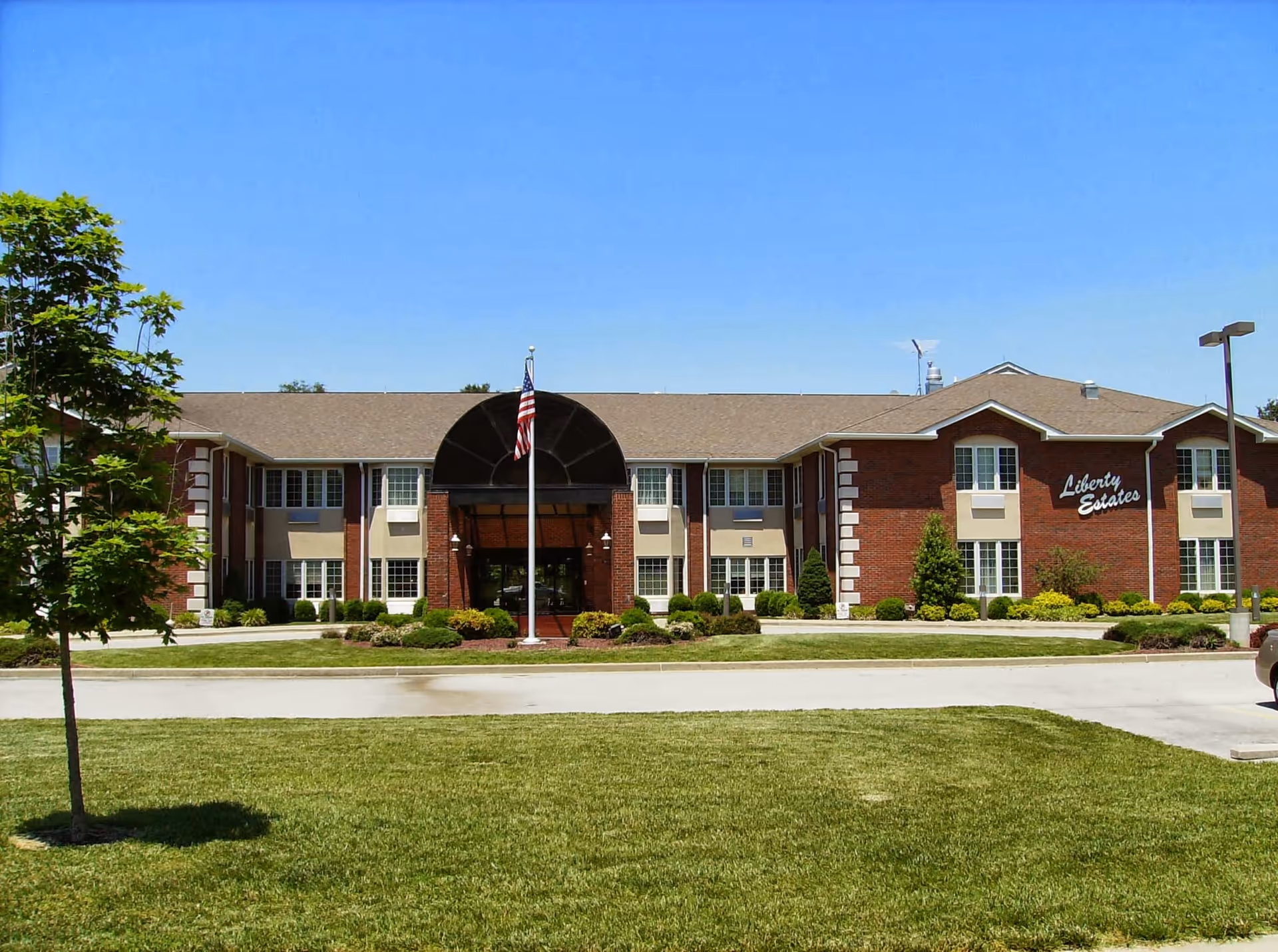 Front exterior view of a two-story brick building with multiple windows, a central entrance with a black awning, an American flag on a flagpole in front, and well-maintained landscaping including grass, bushes, and a tree under a clear blue sky.