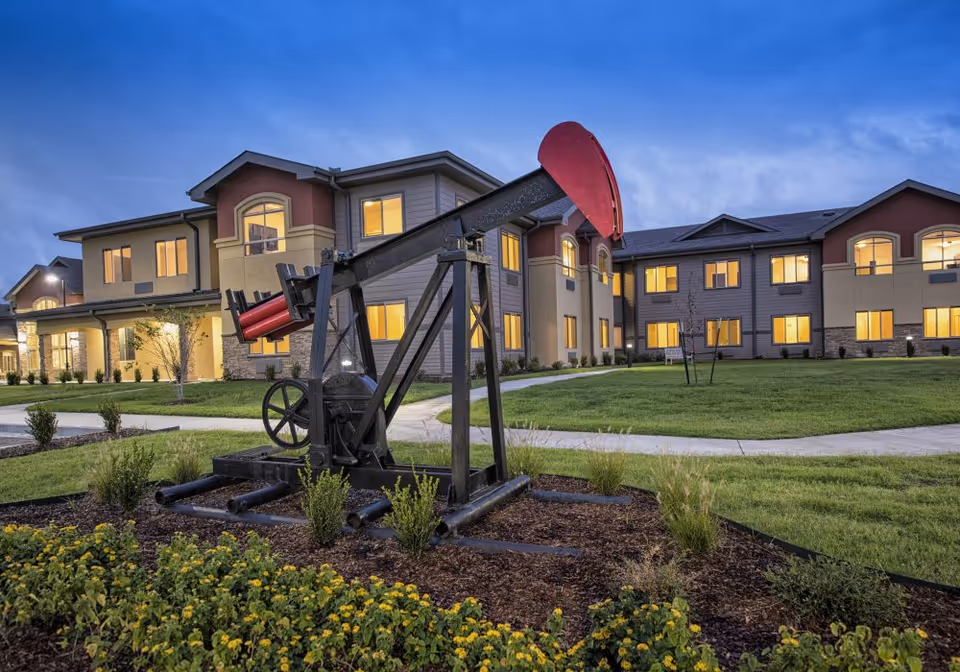 Exterior view of The Stonehaven Assisted Living and Memory Care building at dusk with lights on inside. In the foreground, there is a black and red oil pumpjack sculpture surrounded by landscaping with green grass and yellow flowers.