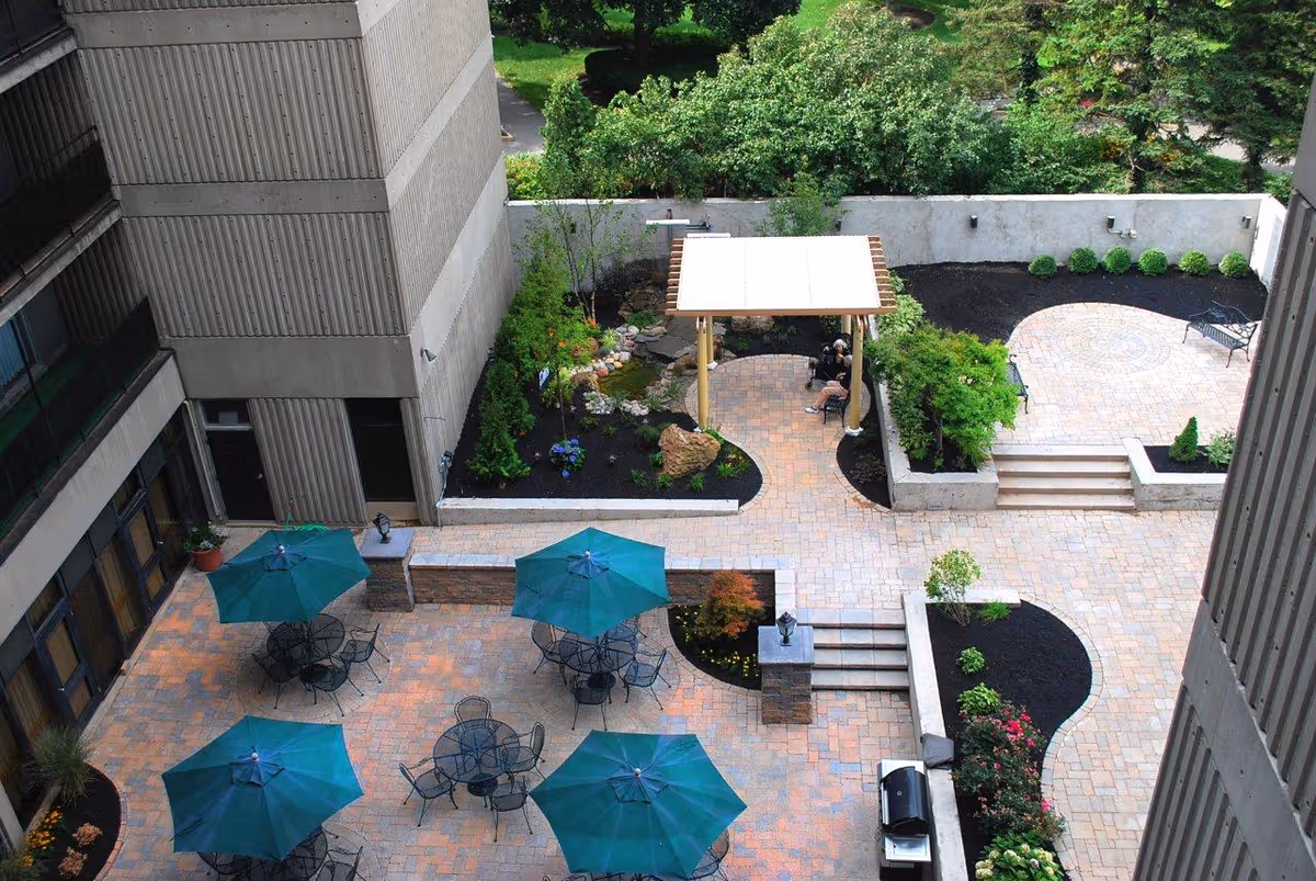 A landscaped outdoor courtyard area with multiple round tables and chairs shaded by green umbrellas. There is a pergola with seating underneath, surrounded by plants and trees. The courtyard is paved with stone tiles and bordered by garden beds with shrubs and flowers. The area is enclosed by tall building walls.