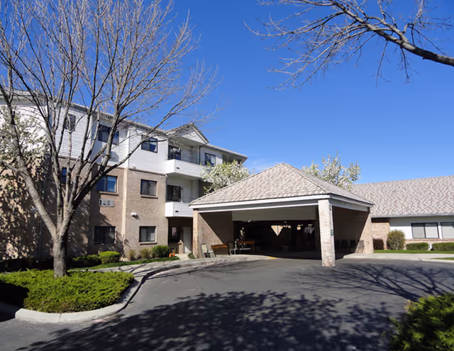 Front entrance and porte-cochère of a multi-story brick senior living building with bare trees and a clear blue sky.