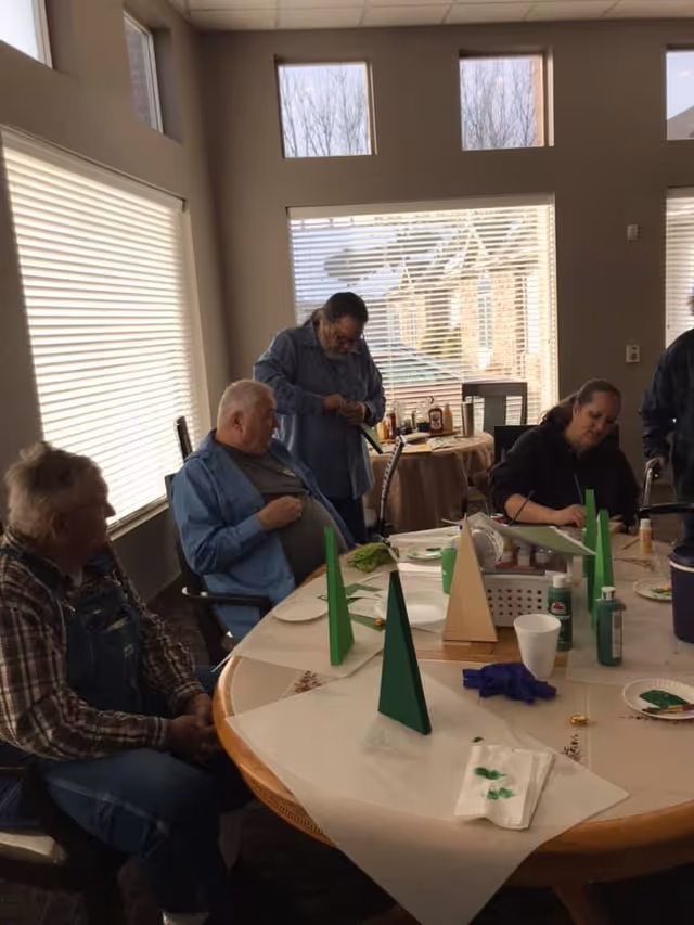 A group of elderly people sitting around a round table in a well-lit room with large windows. The table is covered with a white paper tablecloth and has several green and beige triangular wooden crafts, paint bottles, and painting supplies. One person is standing and appears to be working on a craft, while others are seated and engaged in the activity.