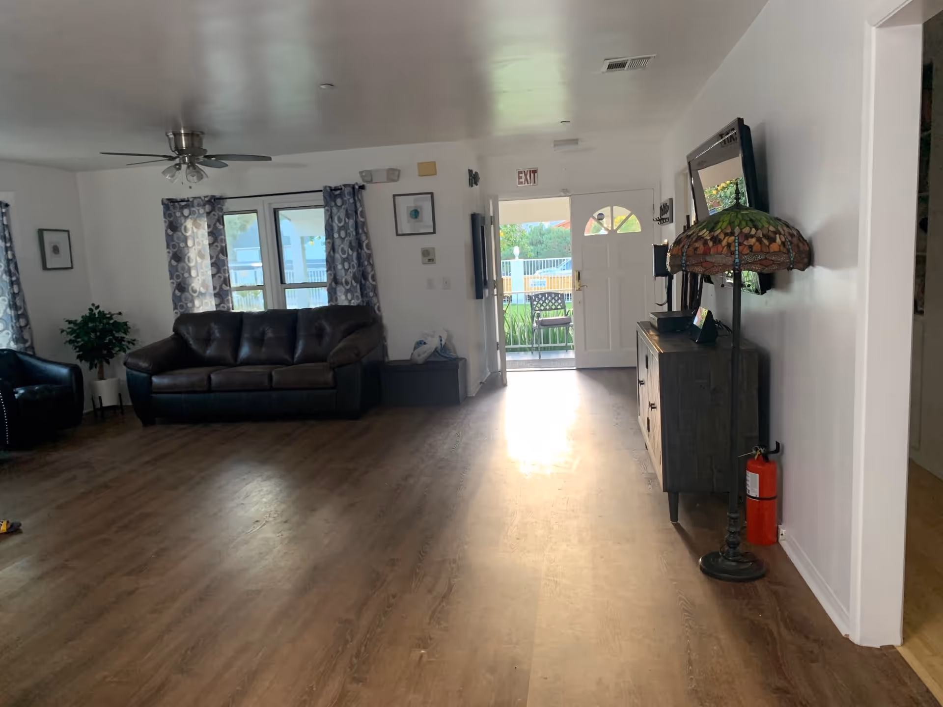 Spacious common living room with a leather sofa and armchair, TV console and Tiffany-style floor lamp, ceiling fan, hardwood floors and an open exit door leading outside.