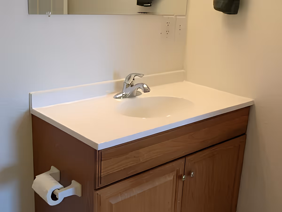 A bathroom vanity with a white countertop and sink, chrome faucet, wooden cabinet, and a toilet paper holder.
