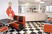 Interior view of a retro-style dining area with black and white checkered floor tiles, red and black cushioned booth seating, and red chairs around tables. A large wall mural of a woman in a red dress is visible on the left wall. The background shows a counter area with kitchen equipment and a window.