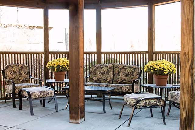 Covered outdoor seating area with cushioned chairs and a loveseat featuring leaf-patterned upholstery, two small tables each holding a pot of yellow flowers, and wooden posts and railing surrounding the space.