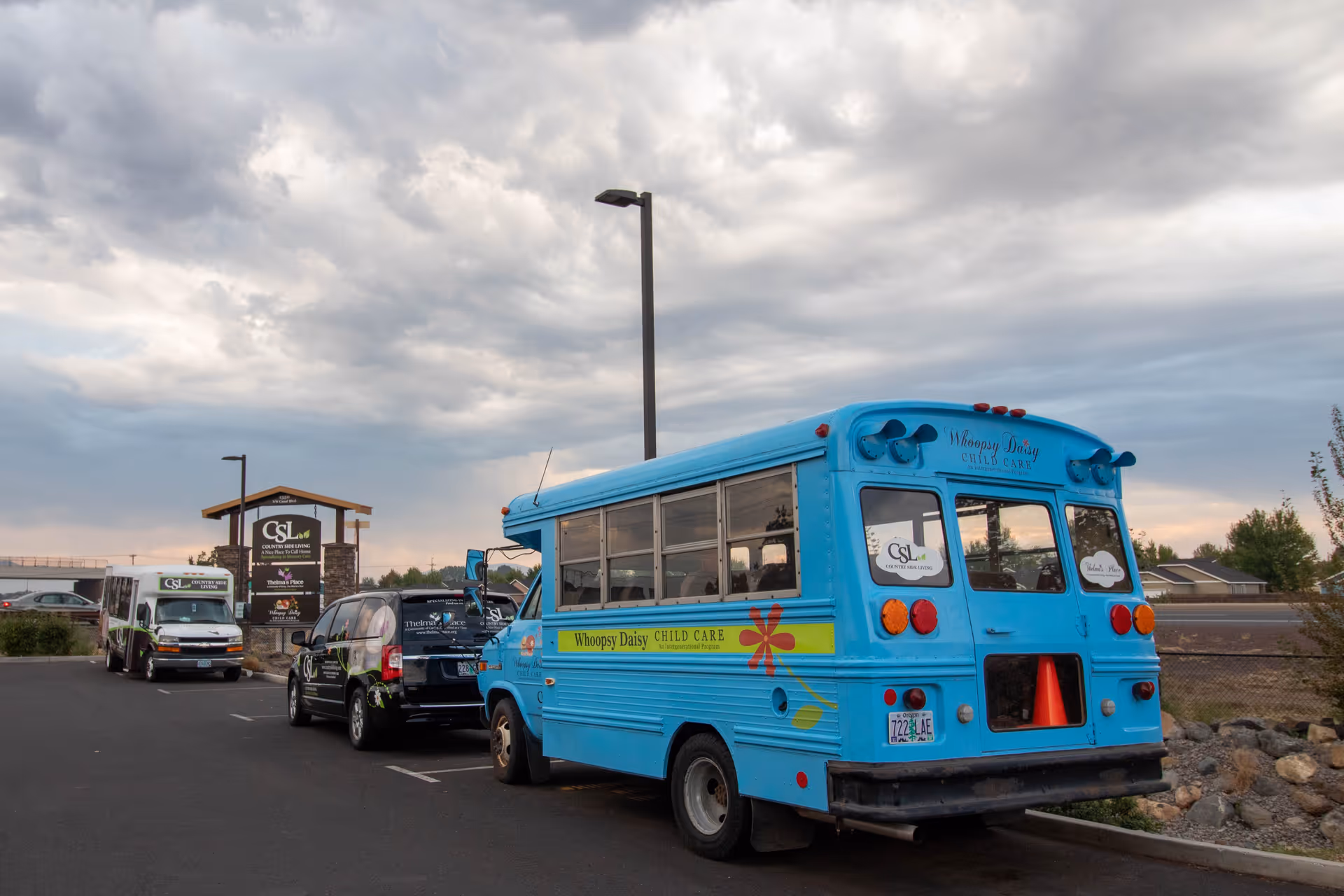 Parking lot with a blue Whoopsy Daisy Child Care bus and two other vehicles parked near a sign for Country Side Living Memory Care facility under a cloudy sky.