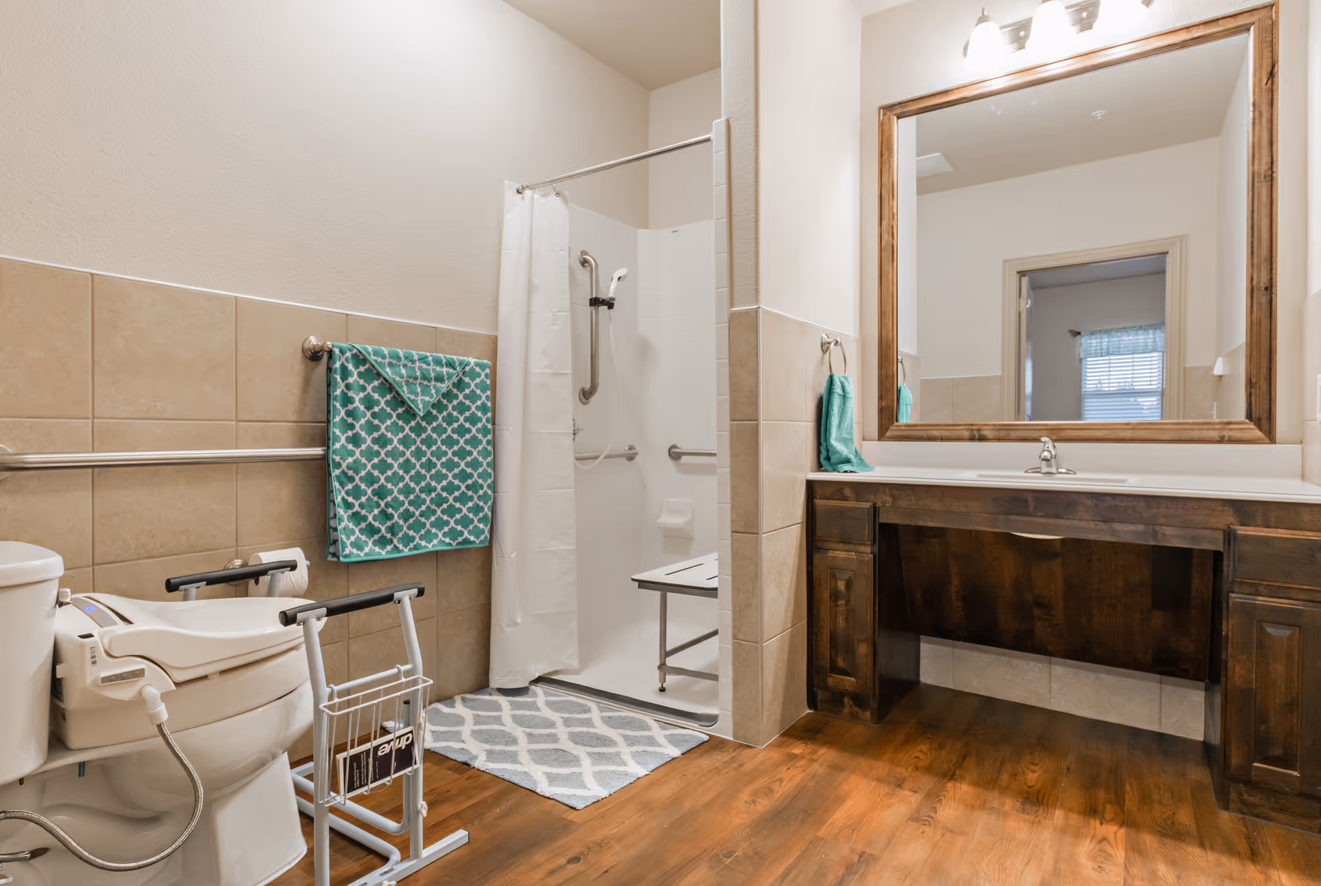 A spacious bathroom with a walk-in shower featuring a white curtain and a shower seat. There is a toilet with safety rails and a bidet attachment. A large wooden framed mirror hangs above a dark wood vanity with a white countertop. The floor is wood, and the walls are partially tiled in beige. A teal towel with a white geometric pattern hangs on a towel bar.