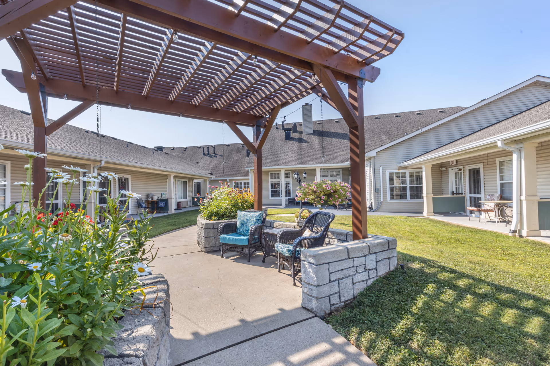 Outdoor seating area at Brookdale Gallatin featuring a wooden pergola with shadows cast on the concrete patio. There are two wicker chairs with blue cushions and a small table between them. Surrounding the patio are stone planters with flowers and greenery. The background shows single-story beige buildings with white trim and windows under a clear blue sky.