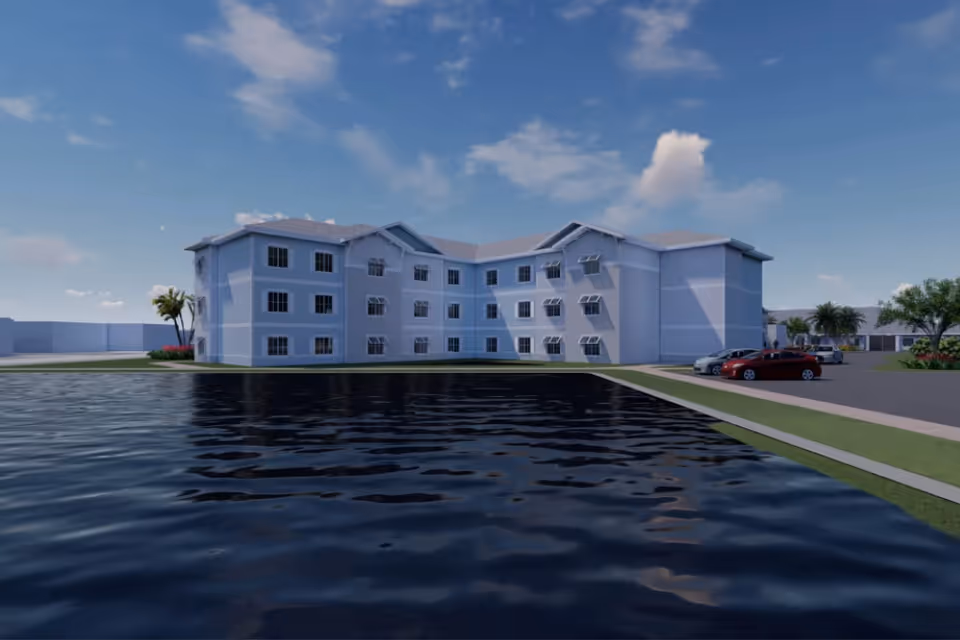 Three-story light-colored senior living building beside a reflective pond with parked cars and palm trees under a blue sky.
