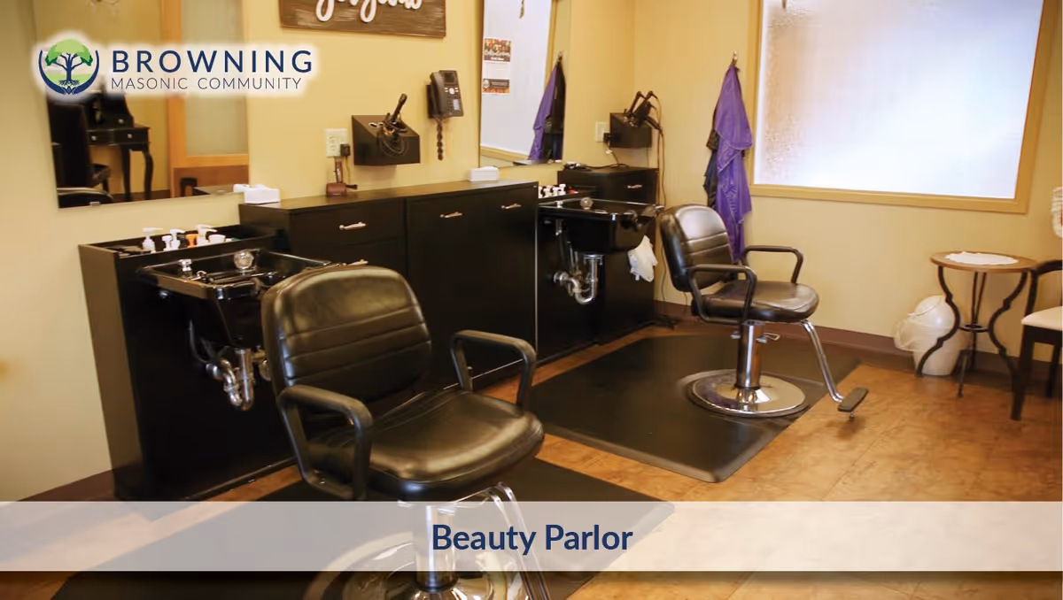 Interior view of a beauty parlor with two black salon chairs in front of black wash basins and cabinets. There is a large mirror on the wall, a telephone mounted on the wall, and a window with frosted glass. A small round table with chairs is visible in the corner.