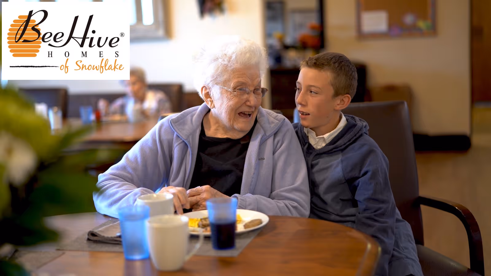 An elderly woman and a young boy sitting together at a dining table in a communal dining area, sharing a joyful moment. The table has plates with food and cups. The background shows other people seated and a bulletin board on the wall.