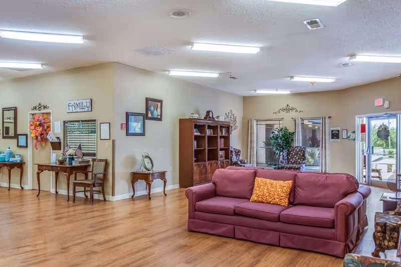A spacious living room area in a nursing and rehabilitation center featuring a maroon sofa with an orange patterned pillow, wooden flooring, a wooden bookshelf, and several framed pictures on the walls. There are two armchairs near a window with curtains, a small table with a clock, and a door leading outside. The room is well-lit with ceiling lights.