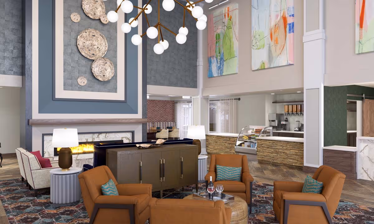 A spacious and modern senior living facility common area with a high ceiling, featuring a seating arrangement of four brown armchairs with teal pillows around a small table. There is a fireplace with decorative wall art above it, two table lamps, and colorful abstract paintings on the walls. In the background, there is a counter area with a display case and beverage dispensers.