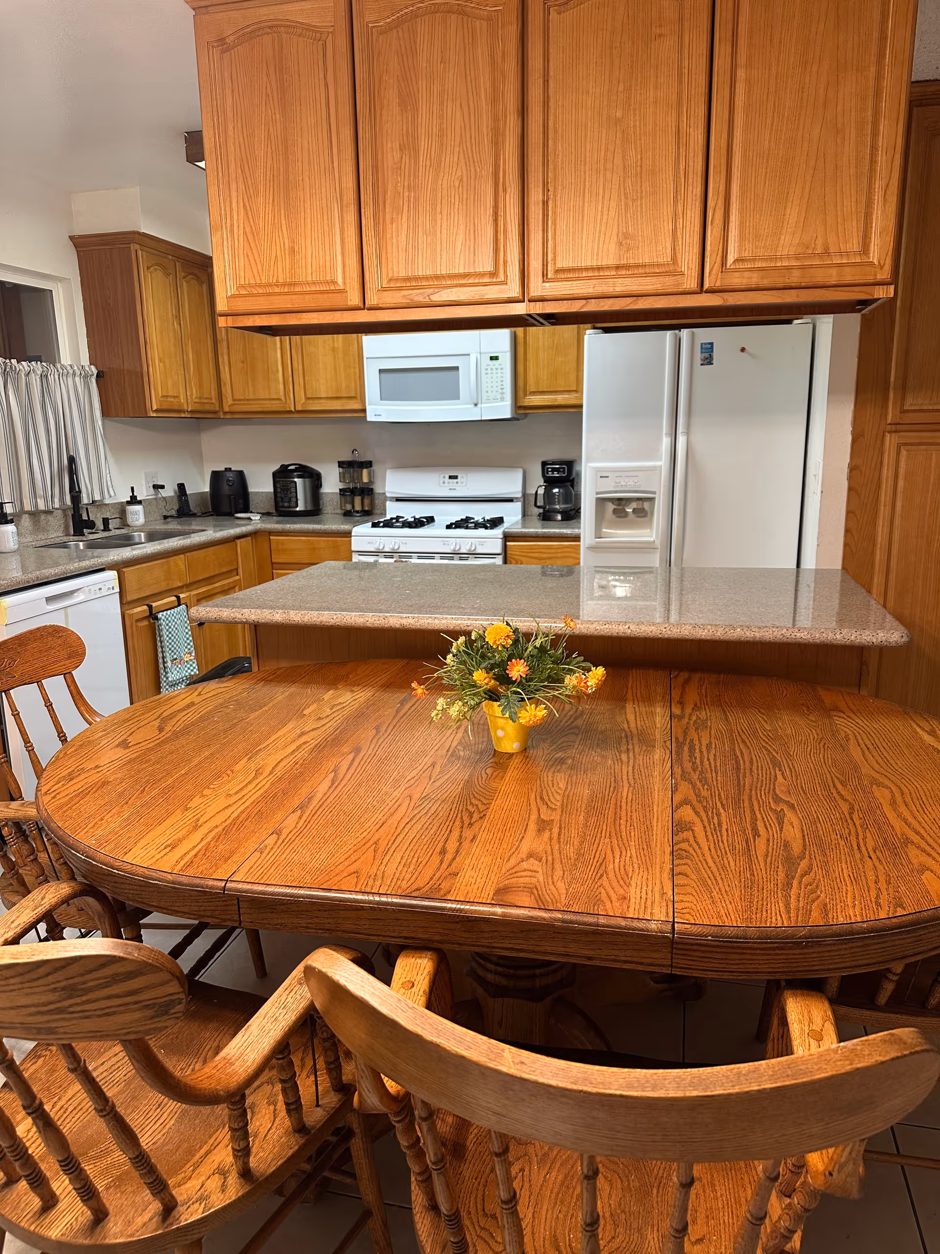 Wooden kitchen with cabinets, fridge, microwave, stove, and a wooden dining table with flowers.