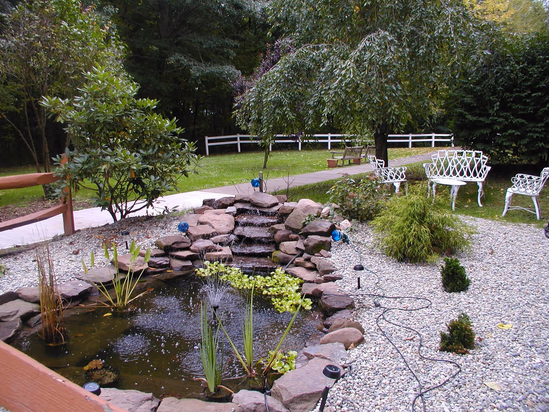 A serene outdoor garden area featuring a small pond with a water fountain and a cascading waterfall made of rocks. Surrounding the pond are various plants and shrubs, white metal chairs and a bench are placed on the grass near a tree. A paved walkway and a white wooden fence are visible in the background with dense trees beyond.