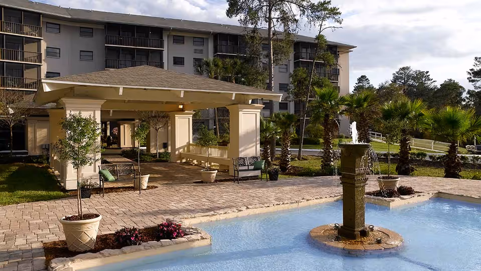 Outdoor area of a senior living facility featuring a covered seating pavilion with benches and chairs, surrounded by potted plants and small trees. In the foreground, there is a decorative water fountain in a shallow pool, with palm trees and a multi-story building in the background under a partly cloudy sky.