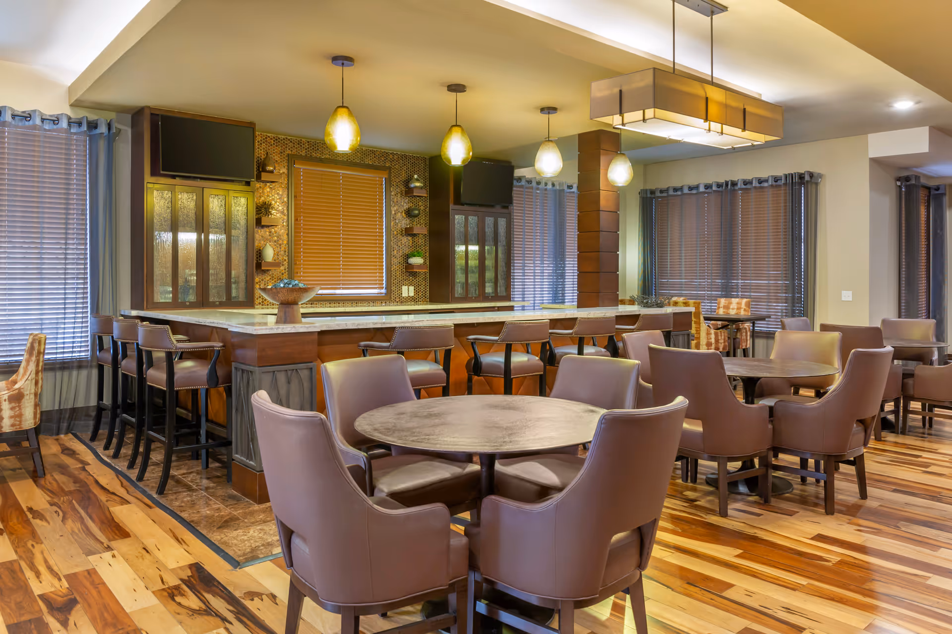 A spacious dining area with multiple round tables surrounded by brown leather chairs. There is a large bar counter with high chairs, pendant lights hanging above, and windows with blinds and curtains. The floor is made of polished wood with a mix of light and dark tones.