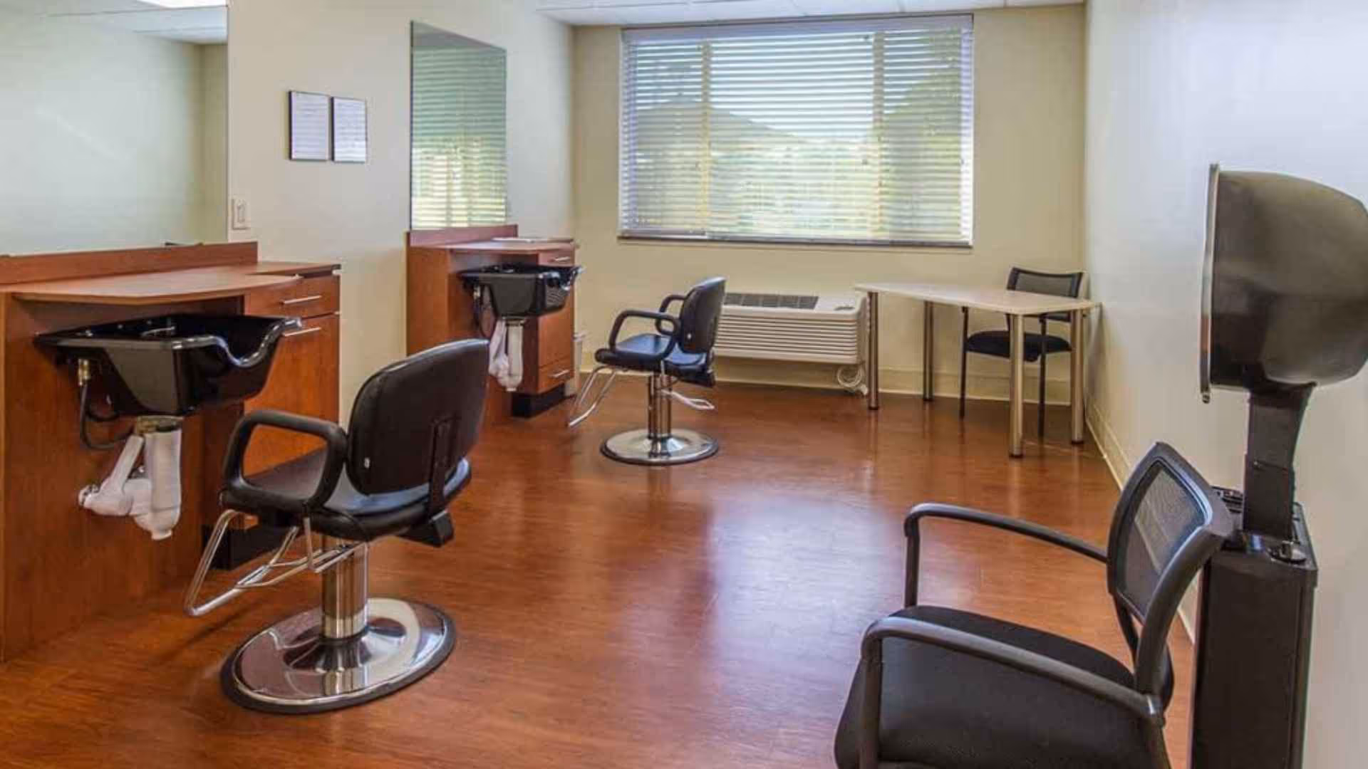 Interior view of a hair salon area in a senior living facility with two black salon chairs in front of wash basins, a large window with blinds, a table with a chair, and a hair dryer chair on the right side.