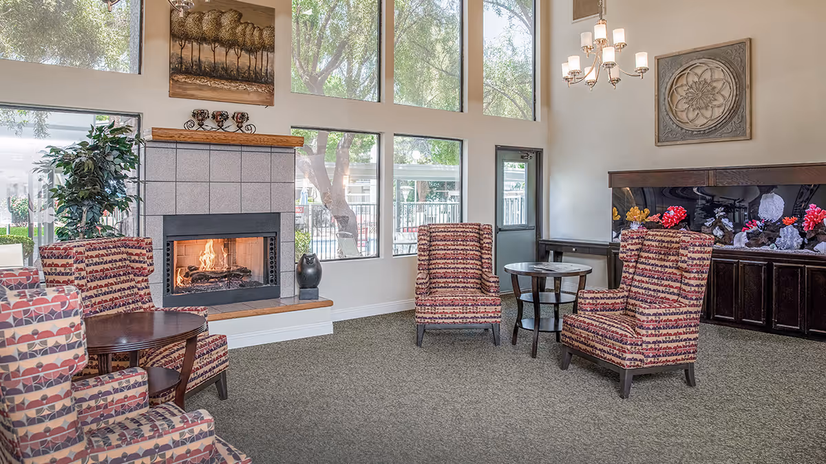 Sunlit common lounge with a lit fireplace, patterned armchairs, side tables, and a large aquarium beneath tall windows.