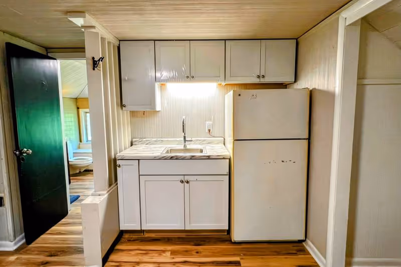Small kitchen area with white cabinets, a marble-patterned countertop, a sink, and a white refrigerator. To the left, an open black door reveals a bathroom with a toilet and window. The floor is wooden and the walls are light-colored.