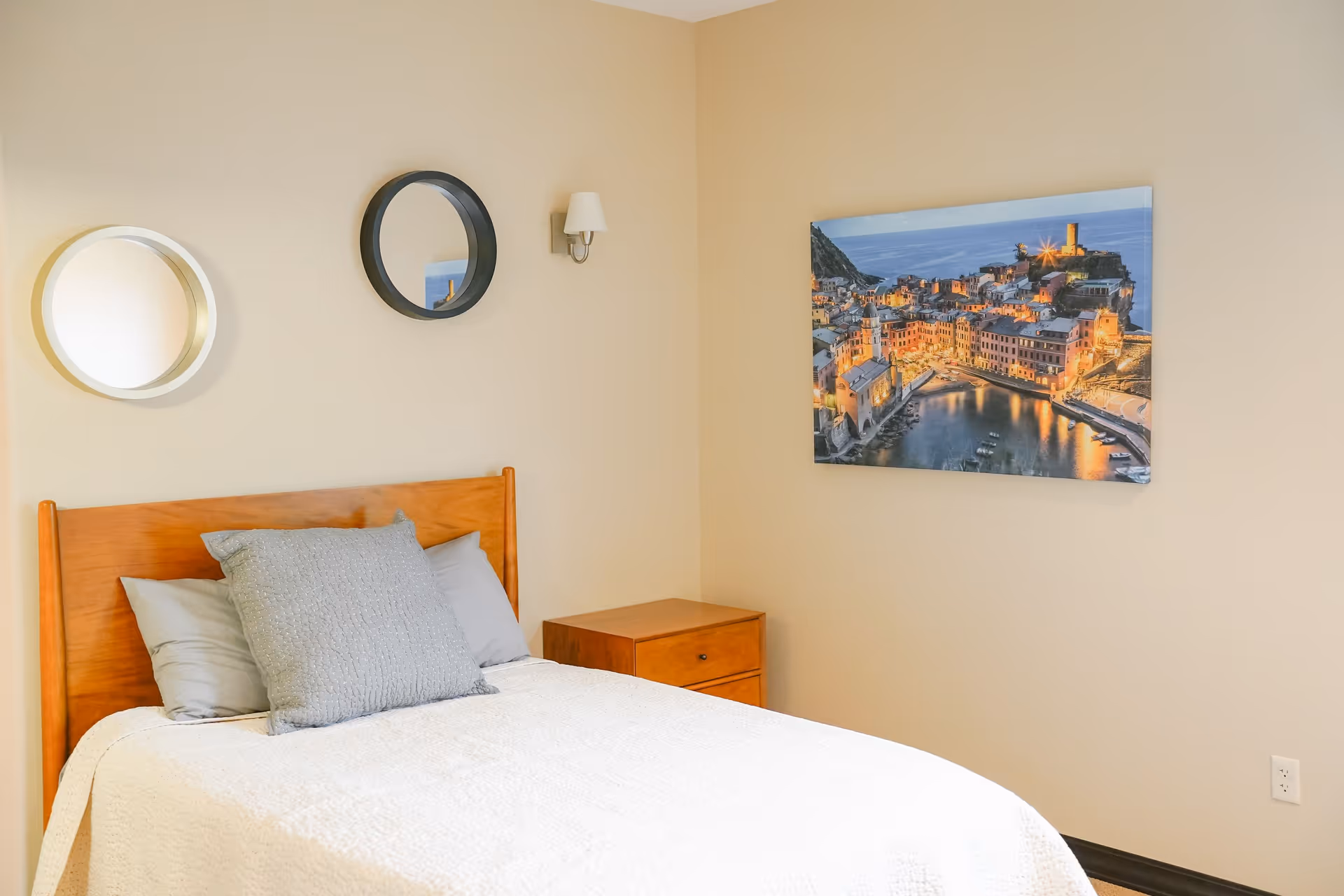 A bedroom with a wooden bed frame, white bedding, and gray pillows. There are two round mirrors and a wall sconce above the bed. A wooden nightstand is beside the bed, and a large framed picture of a coastal town at dusk hangs on the wall.