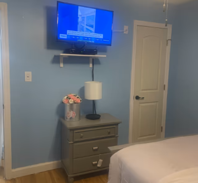 A bedroom with light blue walls featuring a mounted flat-screen TV above a small gray nightstand. On the nightstand, there is a white table lamp and a silver bucket filled with pink and white artificial flowers. A closed white door with a black handle is visible to the right, and part of a bed with white bedding is seen in the foreground.