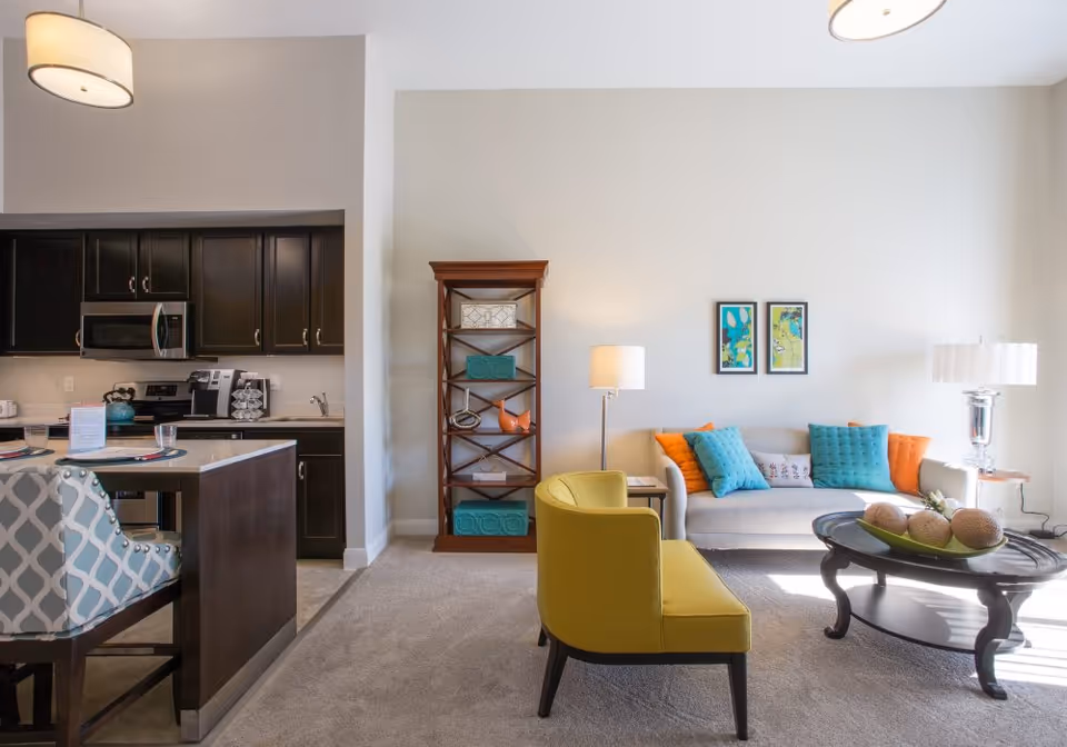 Open-concept living room and kitchen with a beige sofa, colorful throw pillows, a yellow accent chair, round coffee table, and dark wood kitchen cabinets.