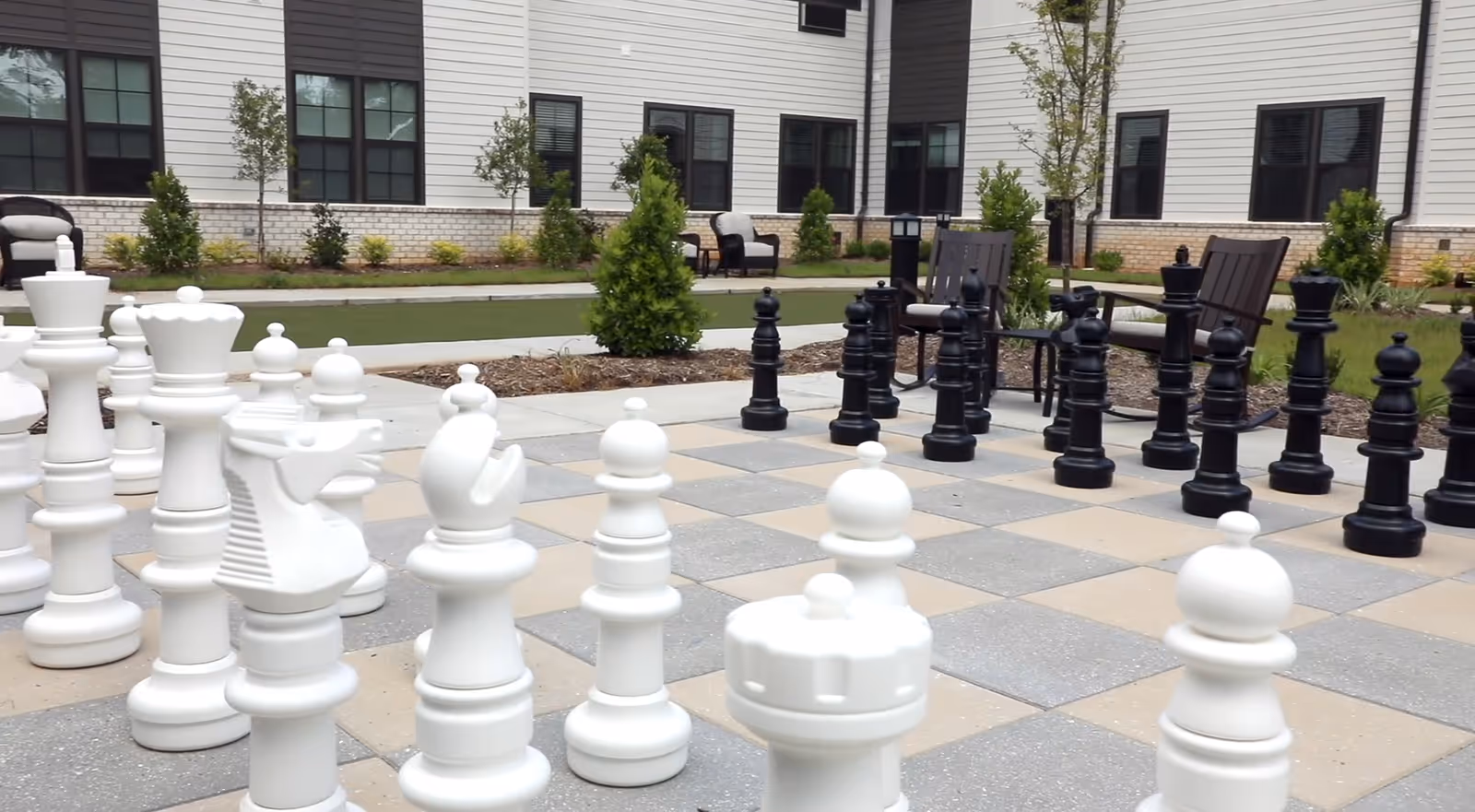 Outdoor area at The Phoenix at Hammond featuring a large chessboard with oversized black and white chess pieces. The background shows a building with multiple windows, some benches, small trees, and landscaped greenery.