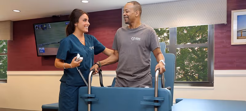 A healthcare worker in blue scrubs assists a male patient using a walker in a rehabilitation room with large windows and a television mounted on the wall.