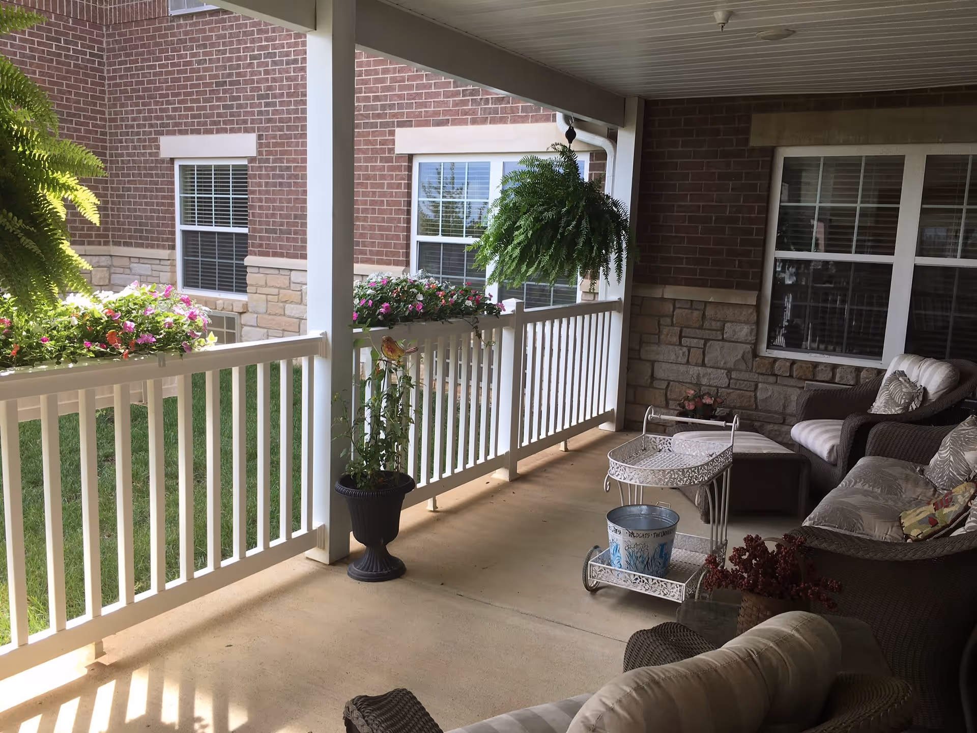 Covered outdoor patio area with white railing, hanging ferns, flower boxes, and potted plants. The patio has wicker furniture with cushions, including chairs and a sofa, and a small white metal table with a bucket on it. The background shows brick and stone exterior walls with windows.