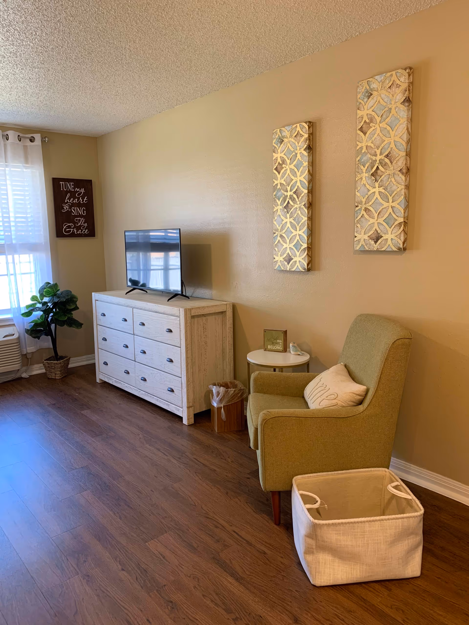A cozy living room area with a green armchair, a small round side table, and a white dresser with a flat-screen TV on top. The walls are beige with two decorative wall hangings and a framed quote near a window with white curtains. There is a potted plant near the window and a white fabric basket on the wooden floor.