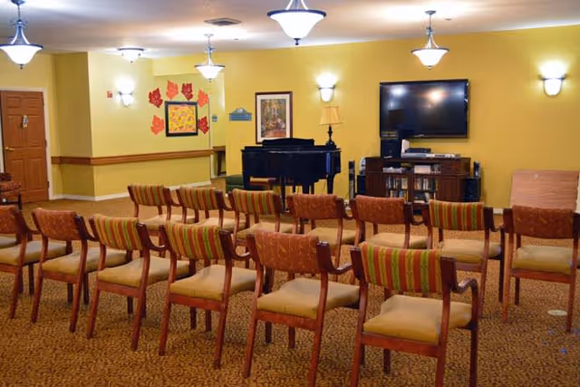 A room with rows of wooden chairs with cushioned seats and backs arranged facing a black grand piano and a large flat-screen TV mounted on a yellow wall. The room has warm lighting from ceiling fixtures and wall sconces, with framed artwork and decorative leaves on the walls.