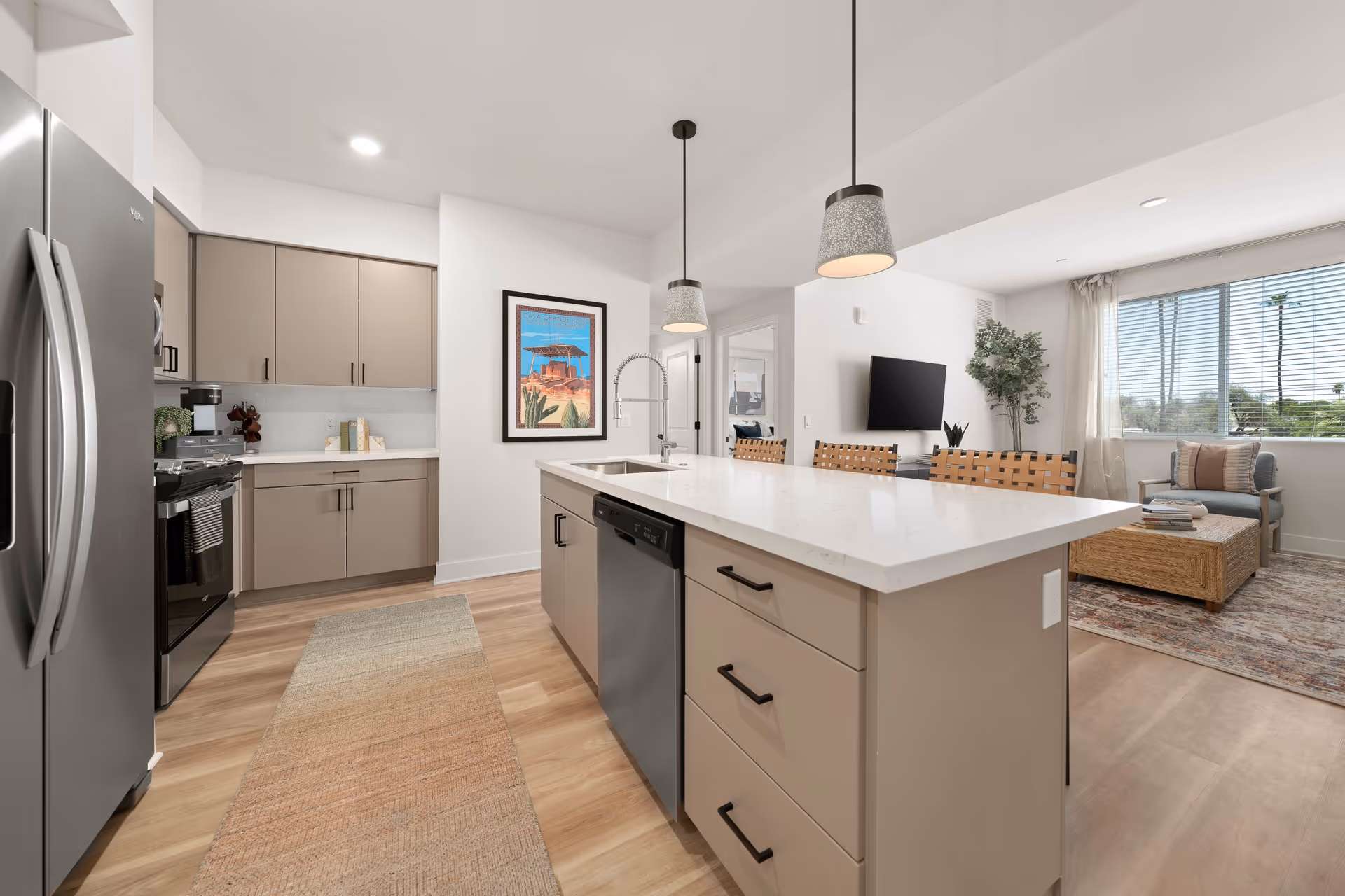 Modern kitchen with beige cabinets, stainless steel refrigerator and dishwasher, a large island with white countertop and three pendant lights above. Adjacent living room area with a window, a wall-mounted TV, a small couch, a coffee table, and a potted plant.