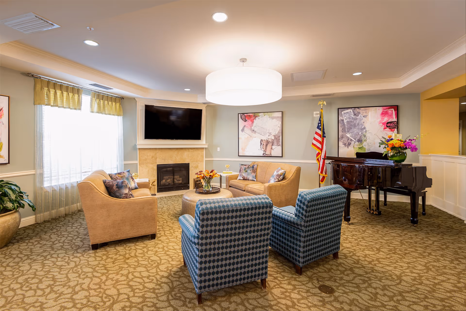 A cozy living room area with two beige sofas and two blue patterned armchairs arranged around a round ottoman with a flower vase on it. There is a fireplace with a mounted flat-screen TV above it, large windows with sheer curtains and valances, abstract artwork on the walls, an American flag, and a black grand piano with a flower arrangement on top. The room has patterned carpet and soft lighting from a large ceiling fixture.