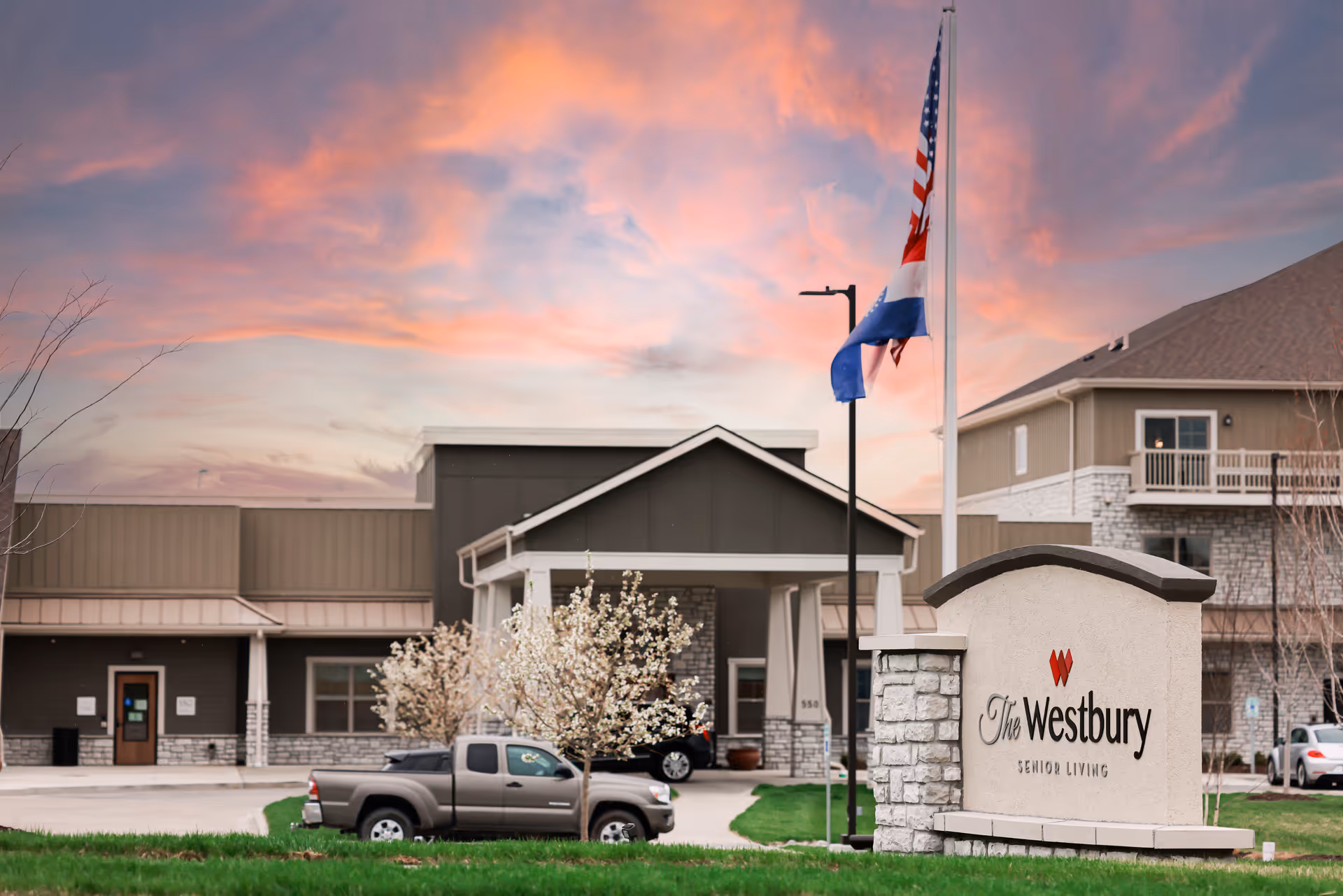 Front exterior of The Westbury Senior Living building with a stone entrance sign, flagpoles and parked vehicles under a colorful sunset sky.