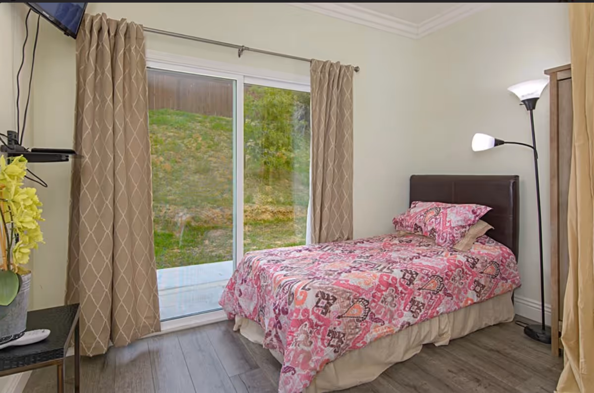 Small bedroom with a single bed in a pink patterned bedspread, sliding glass door with curtains, a floor lamp, and a small side table.