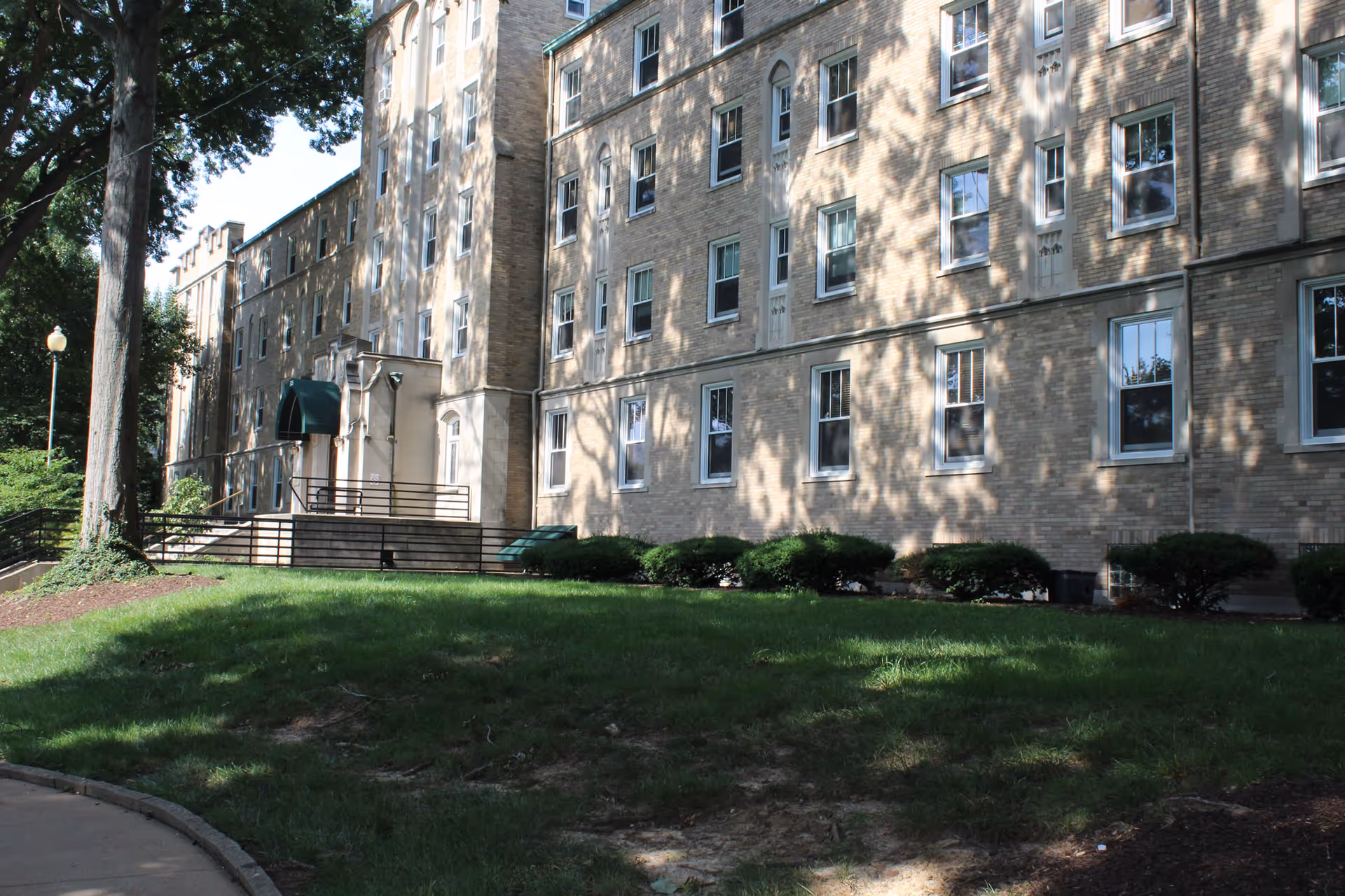 Exterior view of a multi-story brick building with numerous windows, surrounded by green grass, bushes, and large trees casting shadows on the building. A sidewalk curves along the left side of the image.