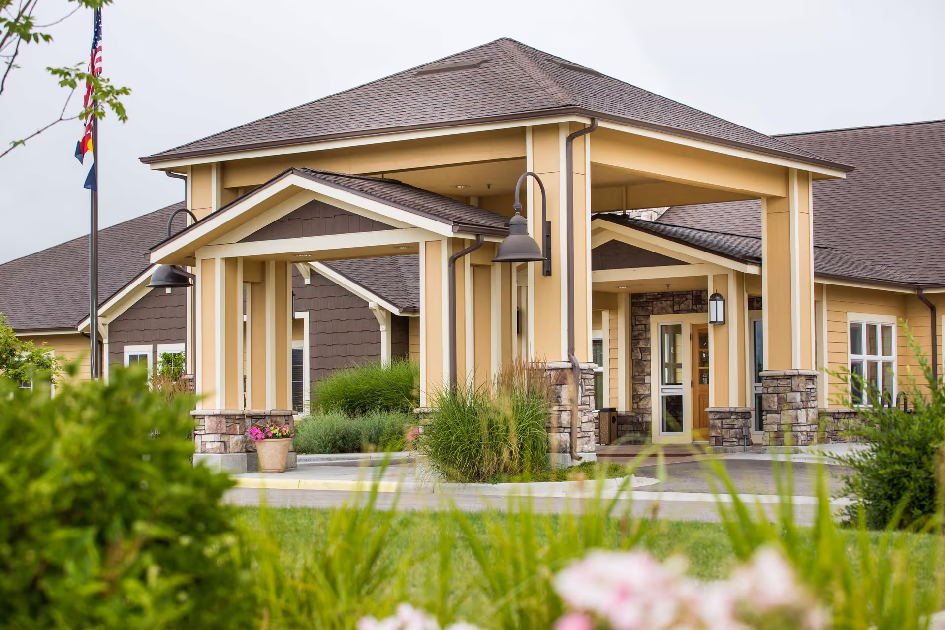 Exterior view of the entrance to Seven Lakes Memory Care facility, featuring a covered drop-off area with beige and brown tones, stone accents on columns, outdoor lighting fixtures, and surrounding greenery including bushes and flowers.