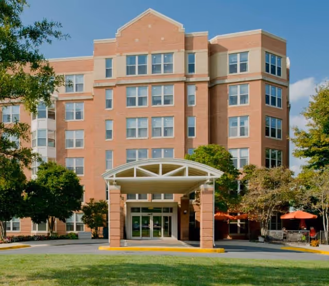 Front exterior view of a multi-story brick building with large windows and a covered entrance, surrounded by trees and outdoor seating with umbrellas under a clear blue sky.