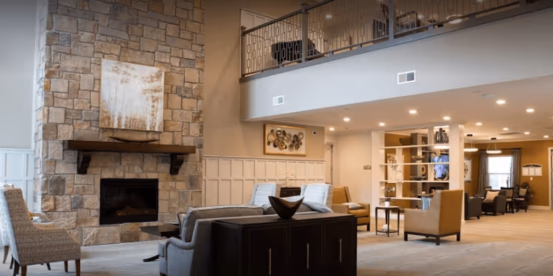 Spacious living room area with a stone fireplace on the left wall, a large painting above the fireplace, and comfortable seating including armchairs and sofas arranged around a central table. The room features a high ceiling with a balcony railing visible above, recessed lighting, and a shelving unit dividing the space. The color scheme is neutral with beige and brown tones.