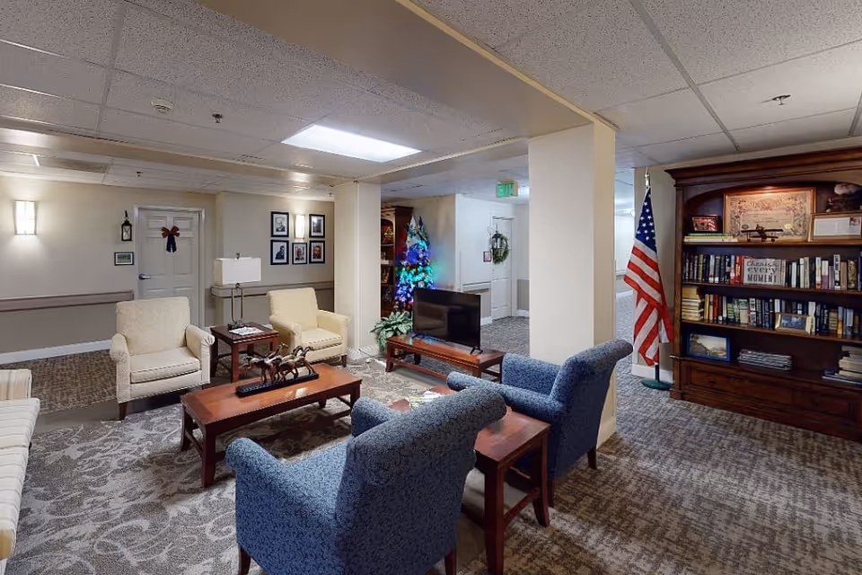 A cozy senior living facility common area with multiple armchairs and a sofa arranged around a wooden coffee table. There is a small TV on a stand, a decorated Christmas tree, an American flag, and a large wooden bookshelf filled with books and framed pictures. The room has carpeted floors, beige walls, and ceiling lights.