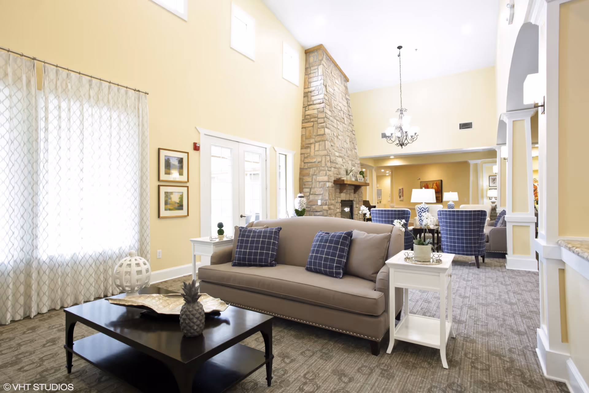 A bright and spacious living room area in an assisted living facility featuring a beige sofa with blue checkered pillows, a black coffee table with decorative items, white side tables with plants, a tall stone fireplace, and several blue upholstered chairs. The room has high ceilings, large windows with sheer curtains, and a chandelier hanging from the ceiling.