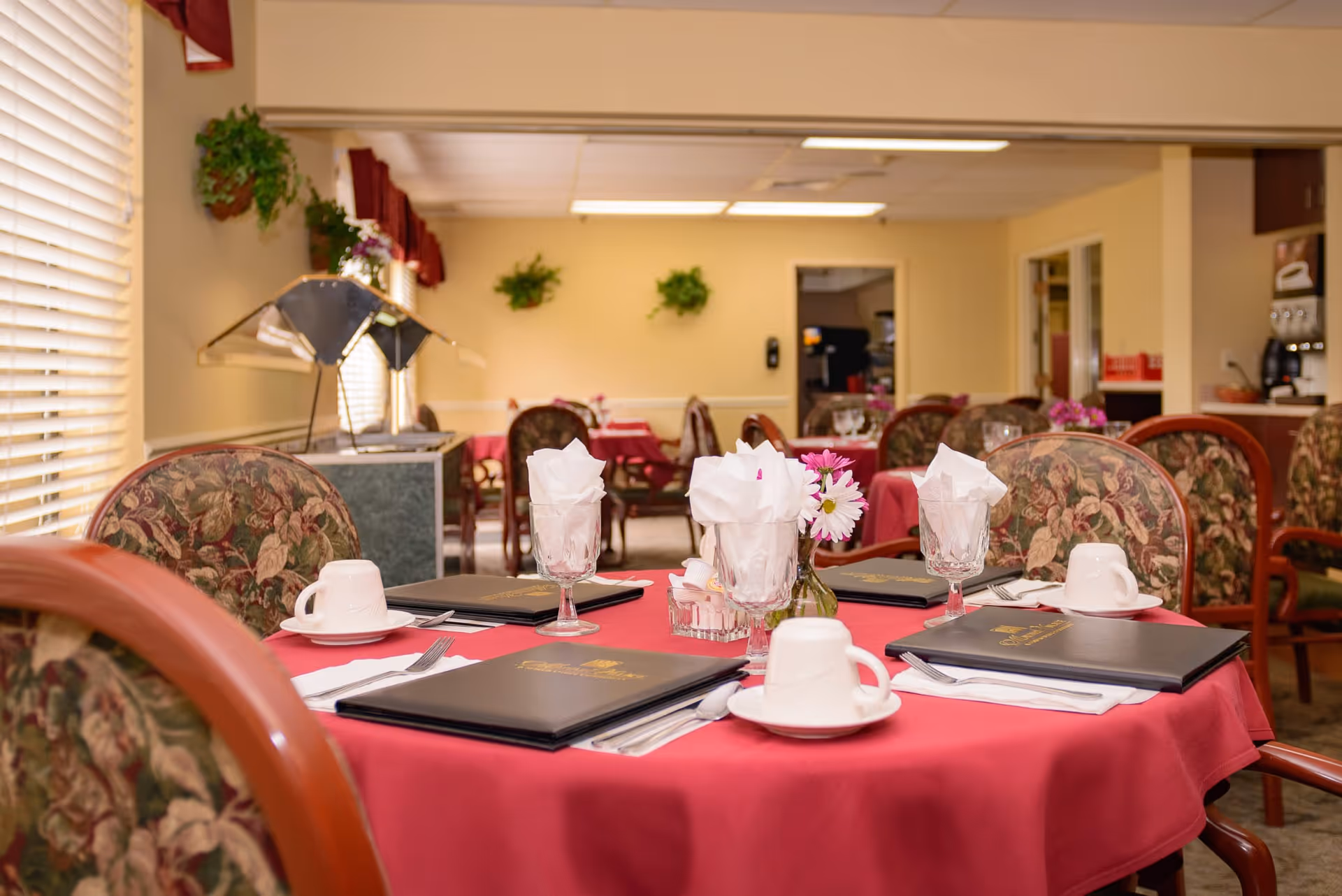 Dining room in Mayfair Village Retirement Center with tables covered in red tablecloths, set with menus, cups, glasses with napkins, and small flower vases. Floral upholstered chairs surround the tables, and there are plants on the walls and windows with blinds letting in natural light.