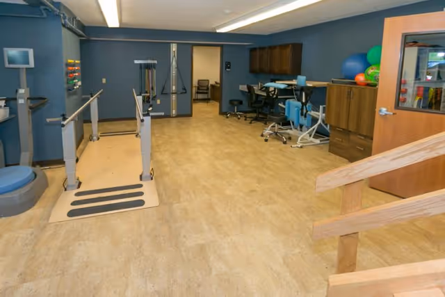 A rehabilitation room with parallel walking bars, exercise equipment, therapy balls, and a practice stair inside a nursing facility.