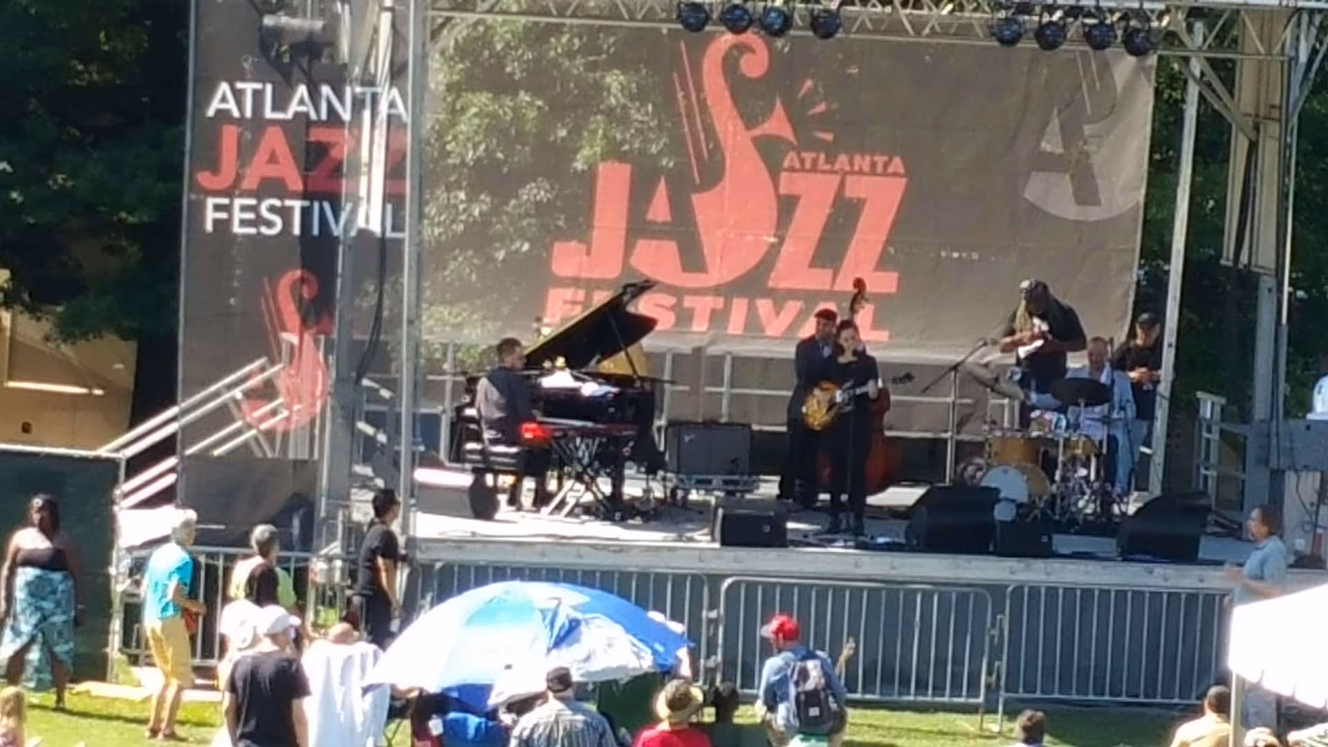 Outdoor stage at the Atlanta Jazz Festival with musicians performing, including a pianist, guitarist, bassist, and drummer. People are gathered in front of the stage, some standing and some sitting under umbrellas, enjoying the live music in a park setting with trees in the background.