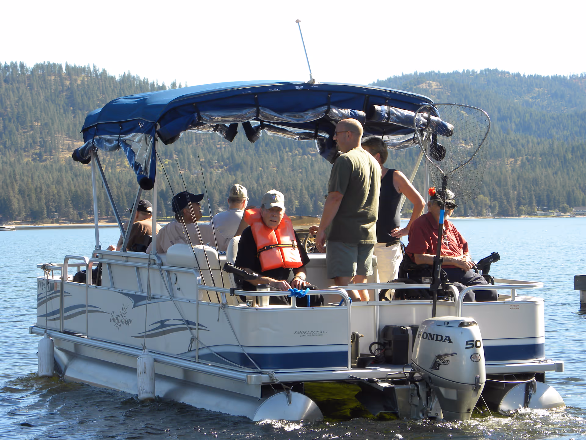 A group of people on a pontoon boat on a lake with forested hills in the background.